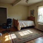 Home workspace merged with personal space, showing an office chair on a kitchen rug among mismatched furniture in a cluttered, shadow-filled room.